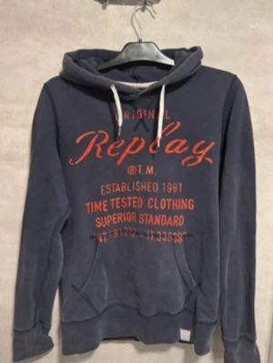 Mörkblå hoodie från Replay - Mörkblå hoodie från Replay med röd textprint på bröstet och vit dragsko i huvan. Klassisk känguruficka framtill och ribbade muddar vid ärmslut och nederkant. Storlek M som passar 170-180cm.