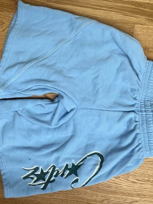 Ljusblå shorts från Croitz - Snygga ljusblå shorts från Croitz med bred resår i midjan och tryckt logga med stjärndetalj på ena benet. Sköna och avslappnade, perfekta för varma dagar. Tillverkade i mjuk bomull för extra komfort.