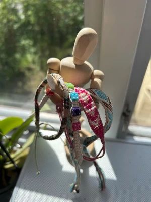 Färgglada vänskapsarmband med pärlor - Säljer flera vänskapsarmband i olika färger som rosa, blå, beige och brunt. Några av armbanden har små färgglada pärlor i form av tärningar. De är flätade och knutna i bomullstråd och passar perfekt för att ge din stil en personlig touch.