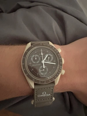 Omega x Swatch Speedmaster klocka - Säljer en stilren Omega x Swatch Speedmaster med grå urtavla och matchande grått band. Klockan har kronograf, tachymeter och tydliga vita visare. Bandet är i syntetmaterial med Omega och Swatch-logga broderad. Perfekt för dig som gillar ikoniska samarbeten.