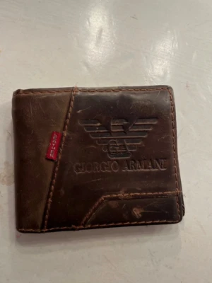 Brun plånbok från Giorgio Armani - Snygg brun plånbok i äkta läder från Giorgio Armani med präglad logga och flera kortfack. Plånboken har en klassisk vikbar design och en röd detalj med texten 'LEATHER' på framsidan. Perfekt för dig som gillar stilrena accessoarer.
