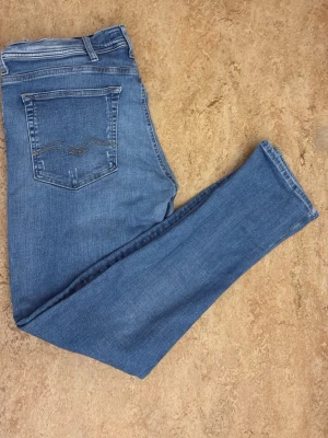 Blå slitna jeans med broderad ficka - Säljer ett par blå jeans med slitningar på framsidan och broderad bakficka. Jeansen har klassisk femficksdesign och raka ben. Materialet är denim och färgen är ljusblå. Perfekt för dig som gillar en avslappnad och trendig look.