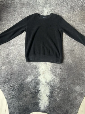 Mörkgrå stickad tröja från Polo Ralph Lauren - Mörkgrå stickad tröja från Polo Ralph Lauren med diskret broderad logga på bröstet. Tröjan har rund halsringning, långa ärmar och är tillverkad i ett mjukt, tunt stickat material som passar perfekt till lager-på-lager-stil.
