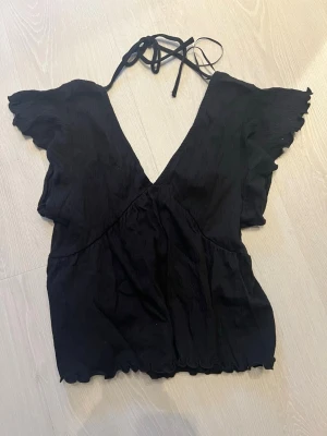 Svart omlottopp med knyt i nacken - Svart topp med djup v-ringning och knyt i nacken. Toppen har korta volangärmar och lätt rynkad nederkant. Perfekt för en trendig och stilren look. Modellen är omlott och ger en feminin siluett.