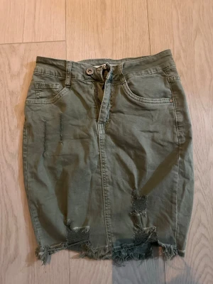 Olivgrön jeanskjol med slitningar - Tuff olivgrön jeanskjol med slitna detaljer och fransig nederkant. Kjolen har klassisk femficksdesign, bälteshällor och dragkedja framtill. Perfekt för dig som gillar en avslappnad och trendig stil.