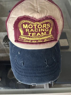 Motors Racing Team keps i bomull - Snygg keps med broderad patch 'Motors Racing Team' framtill. Framsidan är beige med vinröd detalj och text i gult, skärmen är mörkblå med slitna detaljer. Tillverkad i 100% bomull och har en klassisk böjd skärm. Perfekt för en sportig och avslappnad stil.