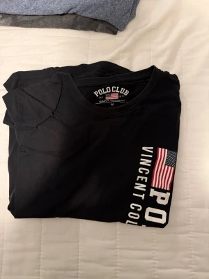 Svart t-shirt från Polo Club - Snygg svart t-shirt från Polo Club med tryck av amerikanska flaggan och texten 'Vincent College' på bröstet. Klassisk rund hals och korta ärmar, perfekt till jeans eller shorts. Enkel men stilren design som funkar till det mesta.