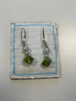 Gröna örhängen med pärlor - Unika örhängen med gröna pärlor och vita pärlor i kombination. Hängena har en cool geometrisk form och små gröna detaljer. Perfekta för dig som vill sticka ut med färg och personlighet. Metallkrokar i silverfärg.