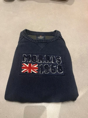 Mörkblå stickad tröja från Morris  - Mörkblå stickad tröja från Morris med broderad text 'Morris 1968' och brittiska flaggan på bröstet. Tröjan har rund halsringning, långa ärmar och ribbade muddar. Tröjan har användes få gånger så är i bra skick. Pris kan diskuteras vid intresse.
