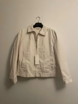 Vit jeansjacka från Zara - Stilren vit jeansjacka från Zara med dragkedja framtill och två stora fickor. Klassisk krage och raka ärmar ger en clean look. Perfekt för dig som gillar minimalistisk stil och vill ha en jacka som funkar till det mesta.