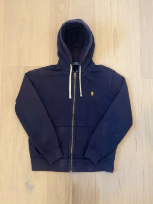 Marinblå hoodie från Polo Ralph Lauren - Säljer en marinblå hoodie från Polo Ralph Lauren med dragkedja, vit dragsko och klassisk gul logga på bröstet. Hoodien har känguruficka och ribbade muddar. Perfekt för en avslappnad och stilren look. Tyvärr så har den en liten liten hål på ärmen, men syns knappt! Fraktar snabbt ✅