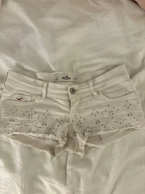Vita broderade shorts från Hollister - Säljer ett par vita shorts från Hollister med broderade detaljer och råa kanter. Shortsen har fem fickor, knappgylf och ett litet Hollister-märke på framsidan. Perfekta för varma dagar och har en fräsch, ungdomlig stil.