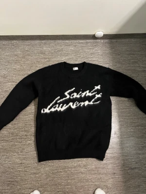 Svart Saint Laurent stickad tröja - Säljer en svart stickad tröja från Saint Laurent med vit logga i graffiti-stil på bröstet. Tröjan har rund halsringning, långa ärmar och ribbade muddar vid ärmslut och nederkant. Perfekt för dig som gillar streetwear och exklusiva märken.