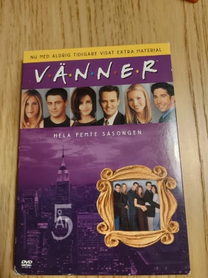 Vänner - Hela femte säsongen - Säljer DVD-boxen 'Vänner - Hela femte säsongen' med extra material. Omslaget är lila med bilder på huvudkaraktärerna och New York. Boxen är tillverkad i kartong och plast, och innehåller fyra DVD-skivor med hela säsongen och bonusavsnitt.