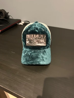 Grön sammetstruckerkeps med tryck - Säljer en unik grön truckerkeps från Goorin Bros/hatstore i sammet med vit mesh baktill. Framtill finns ett svartvitt tryck med motiv och kepsen har justerbar snapback. Perfekt för dig som vill sticka ut med en cool accessoar.