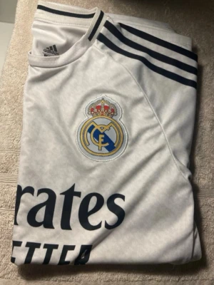 Real Madrid matchtröja Adidas vit Kylian Mbappé - Snygg vit matchtröja från Real Madrid med klubbens broderade emblem på bröstet och mörkblå Adidas-ränder på axlarna. Tillverkad i lätt och ventilerande material, perfekt för fotboll eller träning. Klassisk sportig design med tryck framtill.