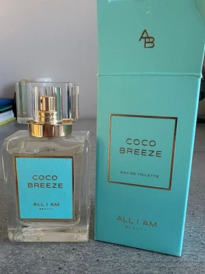 Coco Breeze Eau de Toilette - Fräsch parfym från All I Am Beauty med namnet Coco Breeze. Flaskan är genomskinlig med en stilren, fyrkantig form och har ett turkost etikett med gulddetaljer. Locket är transparent med guldfärgad spraydel. Förpackningen är också turkos med gulddetaljer.