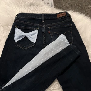 Levi's jeans med blå rosett - Mörkblå jeans från Levi's med unik ljusblå rosett i mesh och pärlor på ena bakfickan. Jeansen har klassisk femficksdesign och raka ben. Insidan av benen har ljusblått tyg med vita prickar som syns när du viker upp dem. Perfekt för dig som vill sticka ut!
