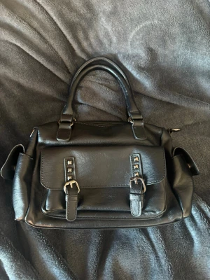 Svart handväska med spännen 🖤🩶 - Snygg svart handväska i fejkläder med dubbla handtag och en stor ficka framtill med två dekorativa spännen. Väskan har sidofickor och dragkedja upptill. Perfekt för dig som gillar en stilren men edgy look.