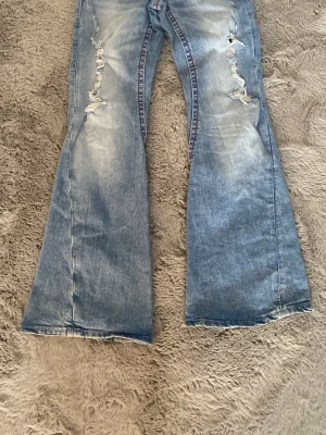 Ljusblå utsvängda jeans med slitningar - Säljer ett par ljusblå jeans med rejält utsvängda ben och slitna detaljer framtill. Jeansen har en tvättad look och dekorativa sömmar längs benen. Perfekta för dig som gillar retrostil och vill sticka ut med din outfit. Pris 300 vanliga priset är 446kr pris går att diskutera tar Swish 