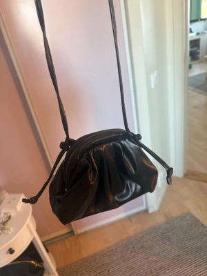 Svart liten axelremsväska i skinn ! - Säljer en svart handväska i mjukt skinn med dragsko och lång axelrem. Väskan har en puffig, veckad design och minimalistisk look som passar perfekt till trendiga outfits. Enkel att bära crossbody eller på axeln.