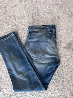 Blå jeans från Replay med tvättad look - Säljer ett par blå jeans från Replay med snyggt tvättad finish och klassiska fem fickor. Jeansen har raka ben, normal midja och detaljerade sömmar på bakfickorna. Märkespatch i brunt läder bak i midjan. Perfekta för en avslappnad stil. W 32 L 30 pris kan diskuteras vid snabb affär. 