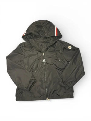 Moncler Windbreaker - Snygg svart vindjacka från Moncler med huva och dragkedja framtill. Jackan har Moncler-logga på ärmen och en rödvit rand på huvan. Tillverkad i lätt och glansigt material som står emot vind. Perfekt för dig som vill ha en stilren och sportig look med fungerande NFC.