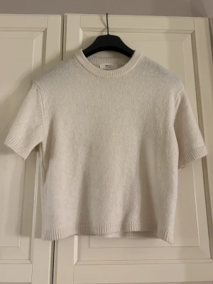 Vit stickad t-shirt från Gina Tricot - Kortärmad stickad t-shirt i vit ullblandning från Gina Tricot. Tröjan har ribbade detaljer vid hals, ärmslut och nederkant. Enkel och stilren design som passar perfekt till många olika outfits.