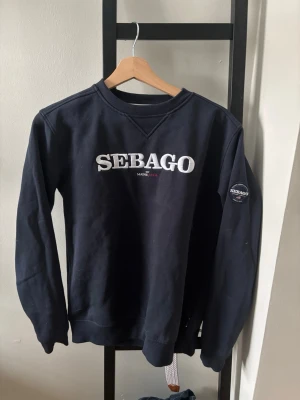 Mörkblå sweatshirt från Sebago - Mörkblå sweatshirt från Sebago med vit broderad logga på bröstet och patch på ärmen. Klassisk rund halsringning och ribbade muddar vid ärmslut och nederkant. Perfekt för en avslappnad och stilren look.