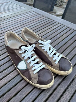 Bruna sneakers från phillipe model - Snygga sneakers i brunt och beige med vita skosnören och en vit patch på sidan. Skorna har en låg profil och en beige sula. Materialet ser ut att vara en mix av mocka och textil, vilket ger en avslappnad och trendig look.