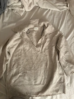 Beige stickad tröja från H&M - Mysig beige stickad tröja från H&M med bred krage och v-ringning. Tröjan har långa ärmar och ribbstickade muddar vid ärmslut och nederkant. Perfekt för lager på lager och enkel att matcha till olika outfits.