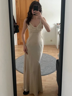 Lång beige klänning - Elegant långklänning i beige/champange med smala axelband. Klänningen har en figurnära passform och öppen rygg. Perfekt för sommarens alla tillfällen. Köpte här på Plick men den var för kort till mig då jag ska ha klackar. Jag är 174cm och bilden är tagen utan skor. 