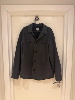 Mörkgrå overshirt från H&M - Stilren mörkgrå overshirt från H&M med klassisk krage och raka ärmar. Två stora bröstfickor framtill och knäppning med svarta knappar. Overshirten är gjord i ett mjukt, tjockare ulliknsnde tyg som ger en avslappnad men snygg look. Perfekt att bära över en t-shirt.                                                         Modellen är 185cm lång och väger 82kg.