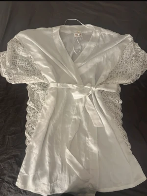 Vit morgonrock med spetsdetaljer - Snygg vit morgonrock med breda spetsärmar och knytband i midjan. Tillverkad i ett lätt tyg med vackra spetsdetaljer längs ärmarna som ger en romantisk och elegant känsla. Perfekt för att chilla hemma eller när du vill känna dig extra fin.