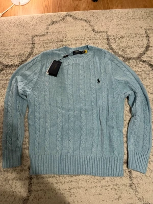 Ljusblå kabelstickad tröja Polo Ralph Lauren - Snygg ljusblå kabelstickad tröja från Polo Ralph Lauren med rund halsringning och klassisk broderad logga på bröstet. Tröjan har ribbade muddar vid ärmslut och nederkant. Perfekt för dig som gillar stilrena och tidlösa plagg. Helt ny aldrig använd 