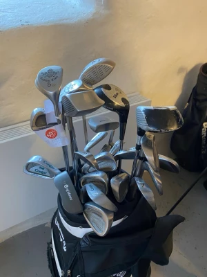 Golfklubbor i bag med flera märken - Säljer ett set golfklubbor i svart och vit bag. Klubborna är av olika märken som Cleveland, Cougar och Custom Star. Setet innehåller järnklubbor, wedgar och drivers i metall och grafit. Perfekt för dig som vill ha ett komplett golfset med blandade klubbor. ALLA KLUBBOR MEN INTE BAGEN!!