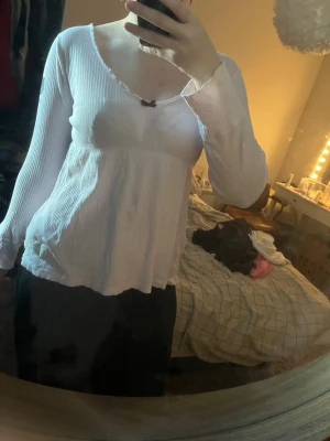 Vit långärmad topp med rosett - Söt vit långärmad topp med ribbad struktur och liten rosett vid urringningen. Toppen har v-ringad hals och är lätt utsvängd nedtill. Perfekt för dig som gillar en enkel men gullig stil.