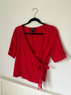 Röd omlottopp från Monki - Snygg röd omlottopp från Monki med knyt i sidan och korta ärmar. Toppen har en djup v-ringning och är gjord i ett lätt, skrynkligt tyg som känns luftigt och bekvämt. Perfekt för dig som vill ha en färgstark och trendig look.