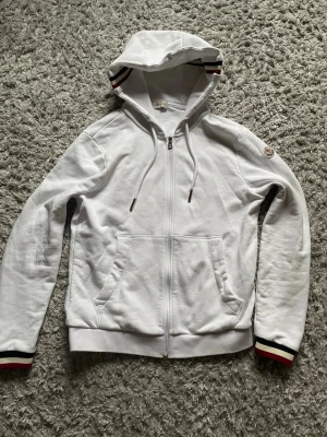 Vit hoodie med dragkedja från Moncler - Snygg vit hoodie från Moncler med dragkedja framtill, huva med dragsko och ribbade muddar med röd-svart-vita ränder. Klassisk Moncler-logga på ärmen och kängurufickor. Perfekt för en clean och sportig look.