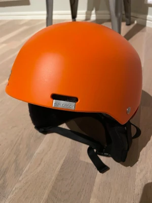 Orange hjälm från Salomon. Jr.  - Säljer en snygg orange hjälm från Salomon, modell JR S. Passar huvudomfång 53-56 cm.  Hjälmen har en slimmad design med svart vaddering på insidan och justerbart hakband. Passar perfekt för cykling eller vintersport. Märke och storlek syns tydligt på hjälmen.
