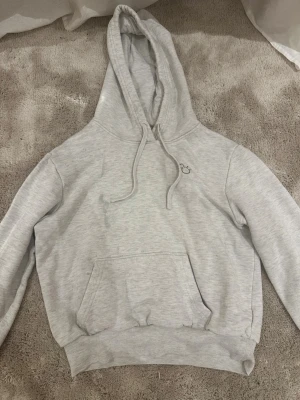 Ljusgrå hoodie med mussepig tryck - Ljusgrå hoodie med huva och dragsnören, samt en stor magficka framtill. Liten broderad detalj på bröstet. Tillverkad i mjuk bomullsblandning och har ribbade muddar vid ärmslut och nederkant. Perfekt för en avslappnad och stilren look. Den har en fläck som inte går bort.