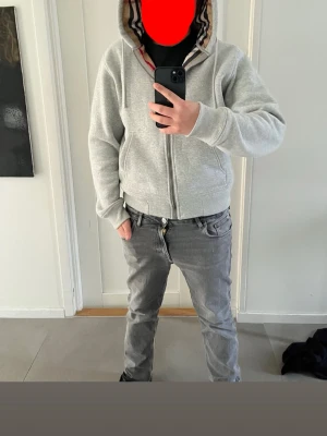 Grå hoodie från Burberry med dragkedja - Säljer en grå hoodie från Burberry med klassiskt rutigt foder i huvan. Hoodien har dragkedja framtill och är tillverkad i mjuk bomull. Perfekt för dig som vill ha en stilren och lyxig look med en twist av ikoniskt mönster. Helt ny!