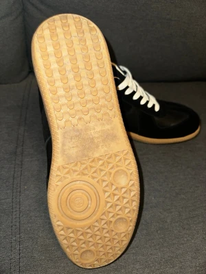 Maison margiala gats black - Stilrena svarta sneakers med vit sula och vita skosnören. Ovandelen är i en mix av mocka och skinn, vilket ger en snygg kontrast. Klassisk låg modell med rund tå och gummisula. Perfekta för dig som gillar en clean och tidlös look.