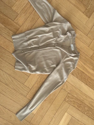 Beige kofta med guldfärgade knappar - Stilren beige kofta från Holly and white  med guldfärgade knappar framtill. Tillverkad i tunt, mjukt material och har långa ärmar samt ribbade muddar. Går inåt längs ner 