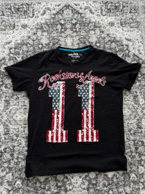 Svart Rockstars & Angels t-shirt - Cool svart t-shirt från Rockstars & Angels med stort tryck av siffran 11 i amerikansk flaggstil och texten 'Rockstars & Angels' över bröstet. T-shirten har rund hals och korta ärmar, perfekt för en avslappnad streetwear-look.