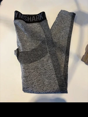 Grå sömlösa leggings från Gymshark - Snygga grå sömlösa leggings från Gymshark med svart resår i midjan där loggan syns tydligt. Leggingsen har mörkare grå detaljer längs sidorna och runt höfterna för en sportig look. Tillverkade i stretchigt material som passar perfekt till träning.
