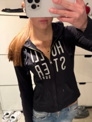 Hollister zip up hoodie - Snygg mörkblå zip up  hoodie från Hollister med dragkedja framtill och stora vita bokstäver på bröstet.                                                                    Storlek S!                                                               Klassisk huva och två fickor framtill. Perfekt till jeans för en avslappnad look.                                          Snöret är lite slitet men annars bra skick!