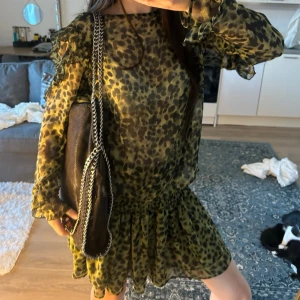 Grön leopardmönstrad klänning - Trendig grön klänning med leopardmönster och volangdetaljer på ärmarna. Klänningen har lång ärm och en lös passform med rynkad midja. Tillverkad i ett lätt, transparent material som ger en luftig känsla.