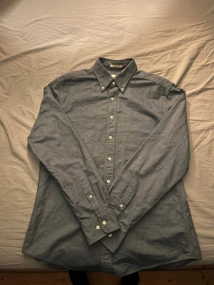 Blå långärmad skjorta med knappar - Klassisk blå skjorta med button-down krage och vita knappar hela vägen framtill. Skjortan har lång ärm och är tillverkad i ett mjukt bomullstyg. Perfekt till både jeans och chinos för en clean look.