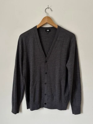 Mörkgrå cardigan från Uniqlo - Stilren mörkgrå cardigan från Uniqlo med v-ringning och knäppning framtill. Tillverkad i tunt, mjukt material som passar perfekt för lager på lager. Långärmad modell med ribbade muddar och diskret design. 8C29